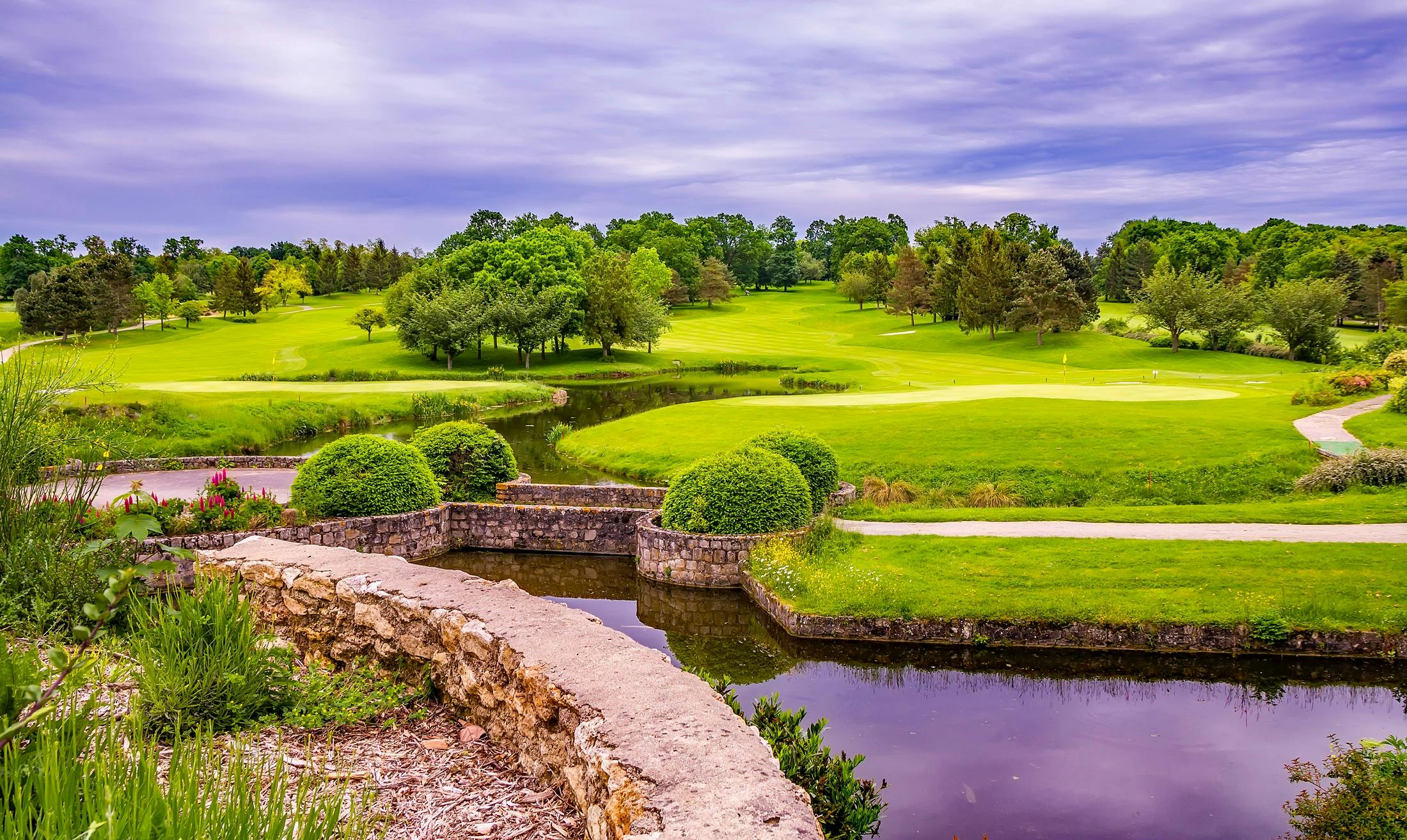 Serene countryside view with lush greenery, pond, and vibrant trees under a cloudy sky.
